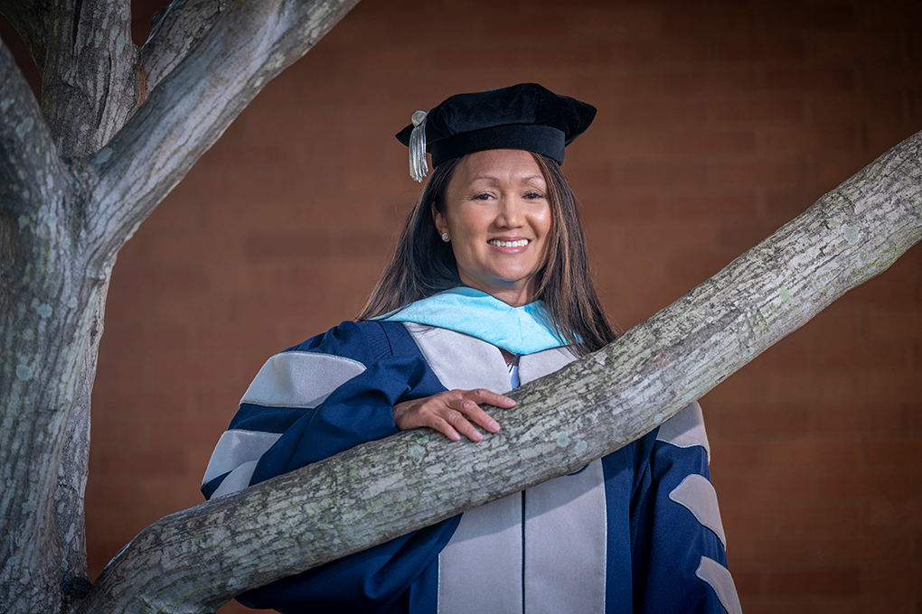A woman wearing graduation regalia stands outside with her hand braced on a tree branch with a brick wall in the background and smiles at the camera.