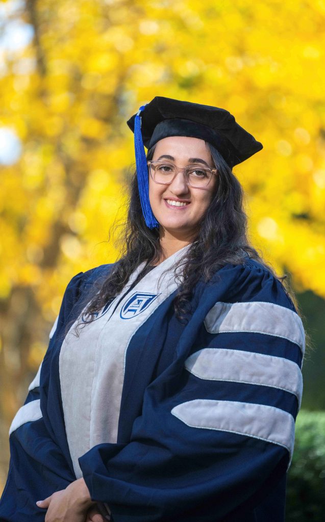 A woman wearing full graduation regalia for a PhD candidate stands outside.