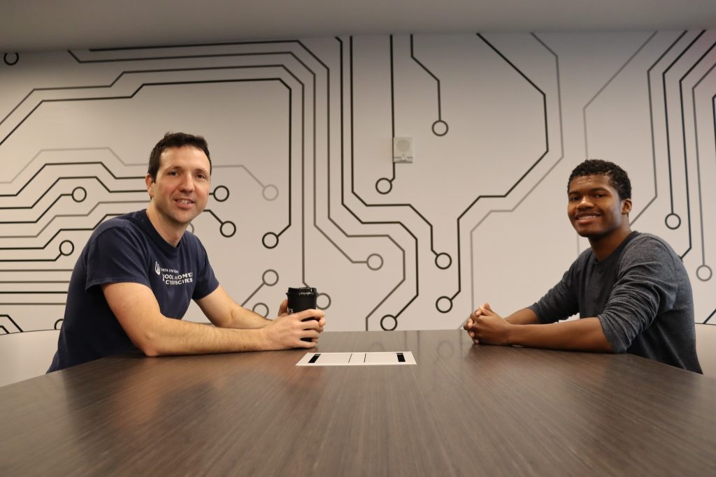 Two individuals sit across from each other at a table, smiling, in a room featuring a wall design that resembles electronic circuitry.