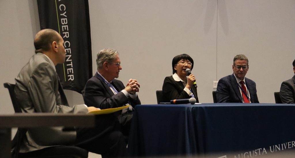 A group of panelists sit at a table during a discussion, with one panelist speaking into a microphone while others listen.