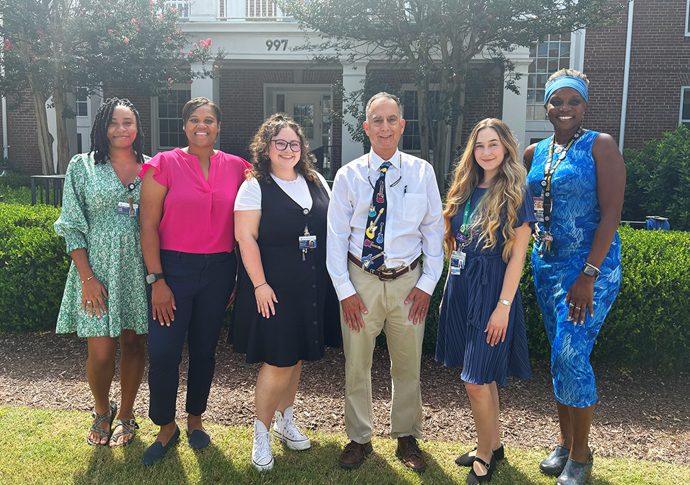 A group of six researchers at a medical college comprised of five women and one man stand outside in front of a building and smile at the camera.