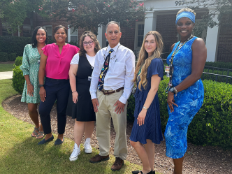 A group of six researchers at a medical college comprised of five women and one man stand outside in front of a building and smile at the camera.