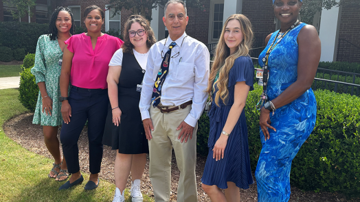 A group of six researchers at a medical college comprised of five women and one man stand outside in front of a building and smile at the camera.