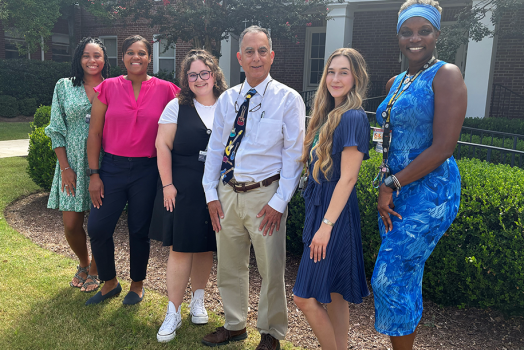A group of six researchers at a medical college comprised of five women and one man stand outside in front of a building and smile at the camera.