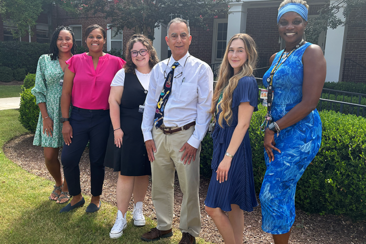 A group of six researchers at a medical college comprised of five women and one man stand outside in front of a building and smile at the camera.