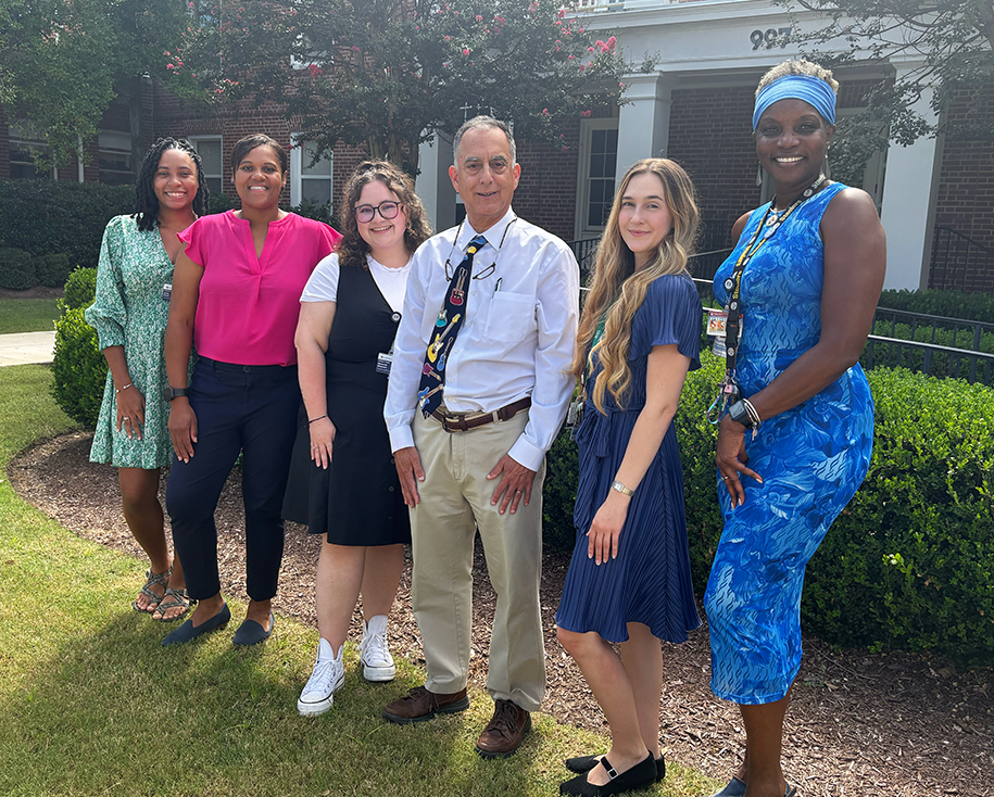 A group of six researchers at a medical college comprised of five women and one man stand outside in front of a building and smile at the camera.