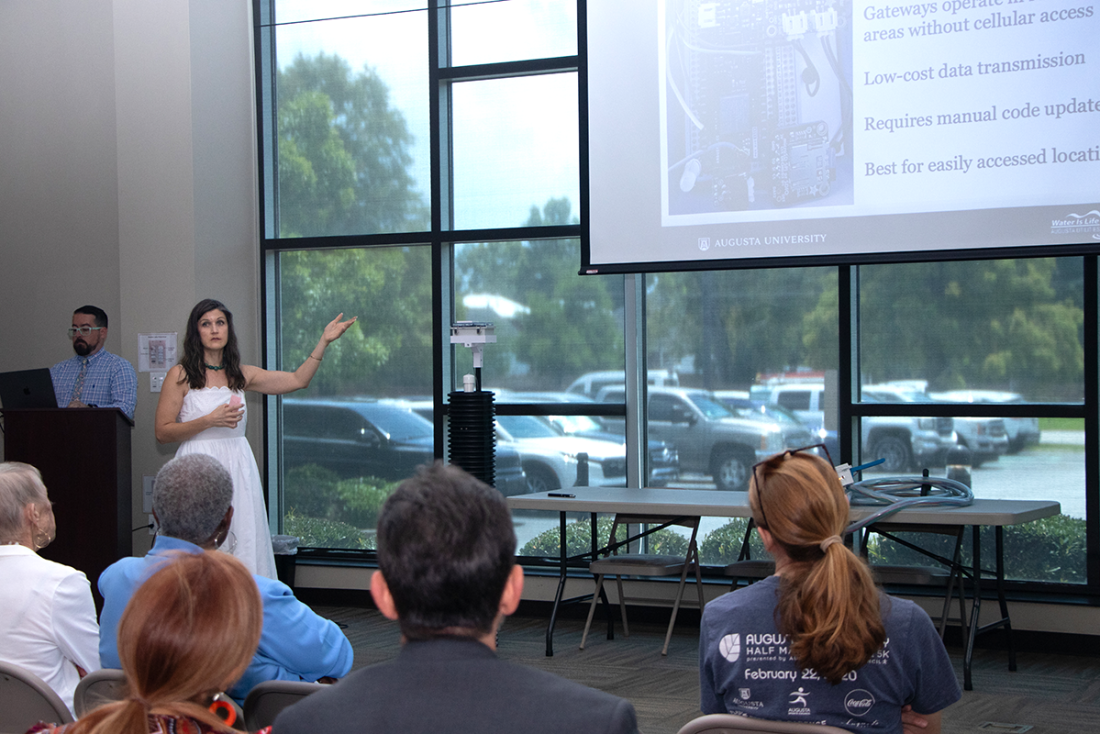 A woman in a white dress, Katy Dunstan, presents a powerpoint of her research to a crowd. Next to her is a six-foot long device made of PVC, which she designed to monitor water levels.