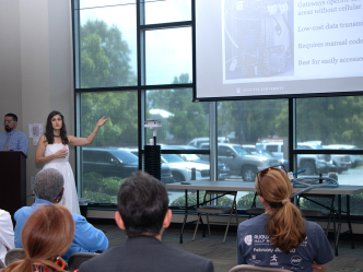 A woman in a white dress, Katy Dunstan, presents a powerpoint of her research to a crowd. Next to her is a six-foot long device made of PVC, which she designed to monitor water levels.