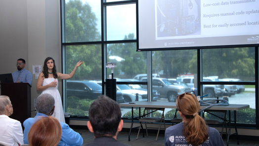 A woman in a white dress, Katy Dunstan, presents a powerpoint of her research to a crowd. Next to her is a six-foot long device made of PVC, which she designed to monitor water levels.