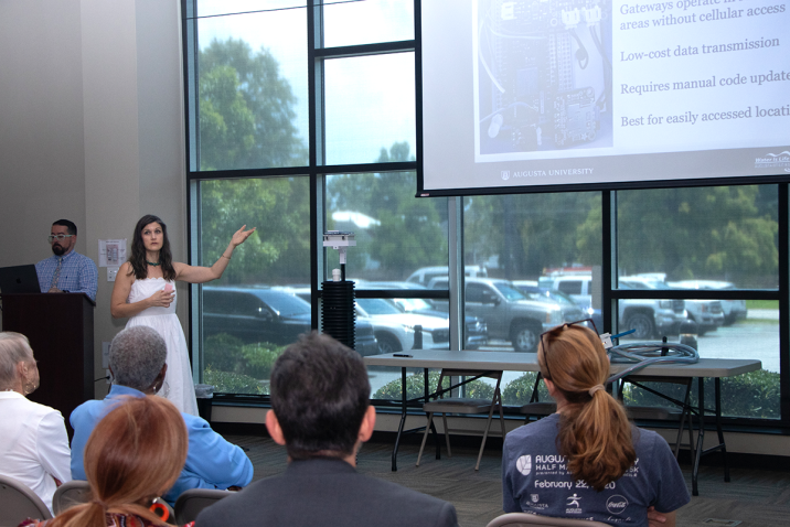 A woman in a white dress, Katy Dunstan, presents a powerpoint of her research to a crowd. Next to her is a six-foot long device made of PVC, which she designed to monitor water levels.
