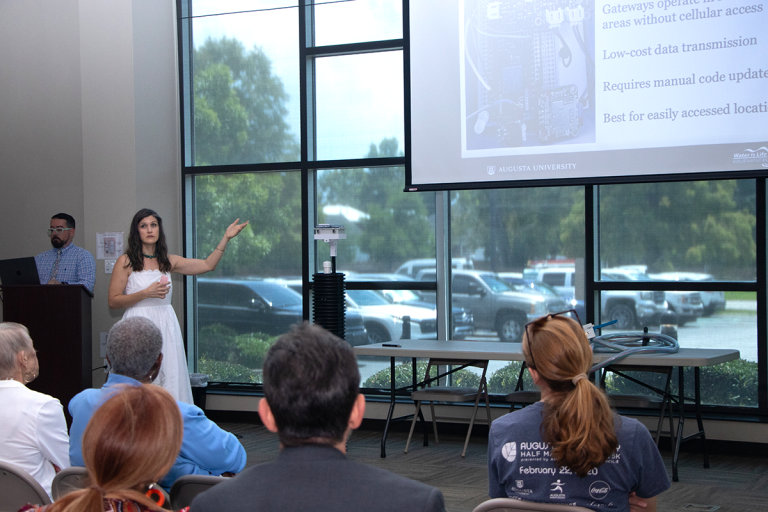 A woman in a white dress, Katy Dunstan, presents a powerpoint of her research to a crowd. Next to her is a six-foot long device made of PVC, which she designed to monitor water levels.