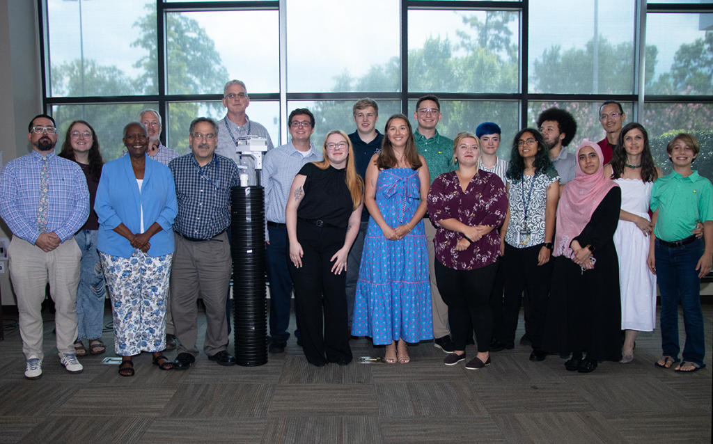 A group stands around a six-foot tall PVC pipe device used to measure water depth.