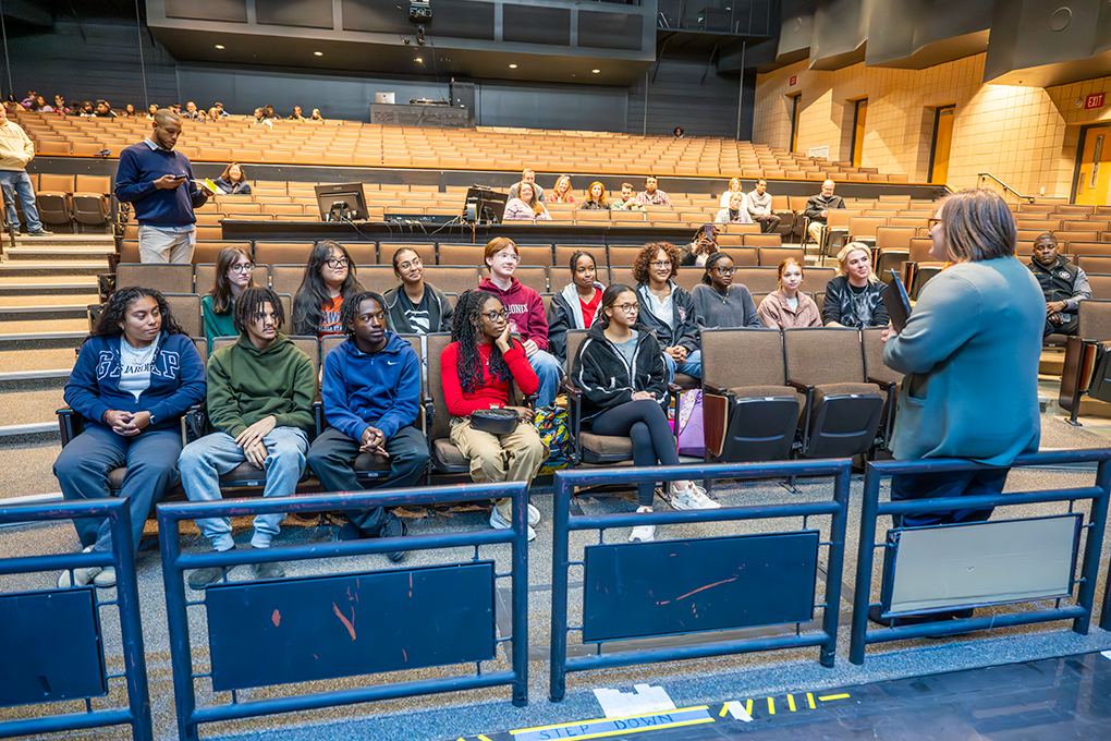 A group of high school students sit in an auditorium and listen to a university faculty member.