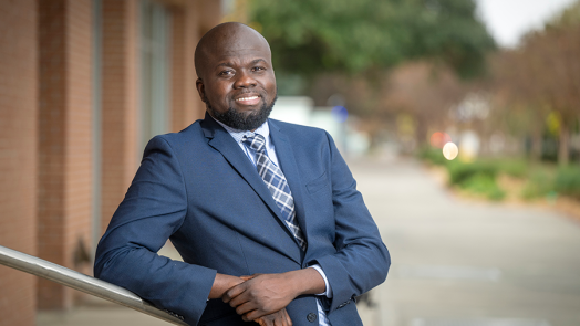 A man in a suit and tie leans against a railing outside of a modern research building on a college campus.