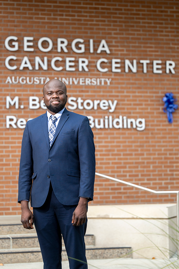 A man in a suit stands outside of a large, modern research building on a college campus. There is a large sign on the wall behind him that reads, "Georgia Cancer Center. M. Bert Story Research Building."