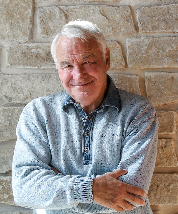 An older man smiles while leans against a stone wall with his arms crossed.