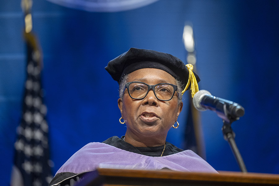 woman with glasses wearing a graduation cap and gown speaking at a podium. 