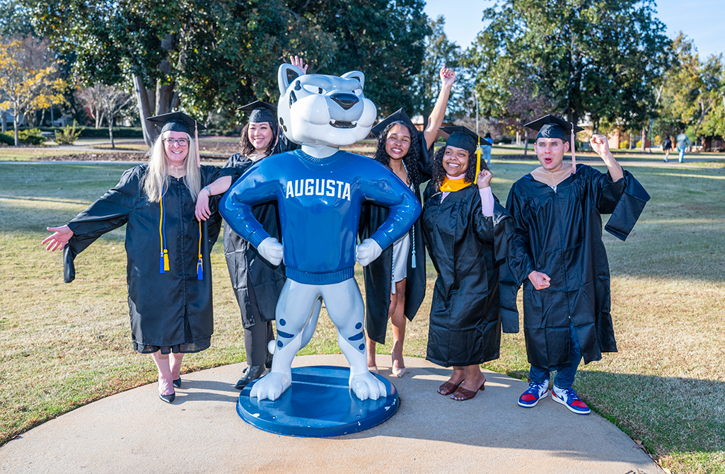 Four women and one man pose with large cat statue wearing graduation caps and gowns.
