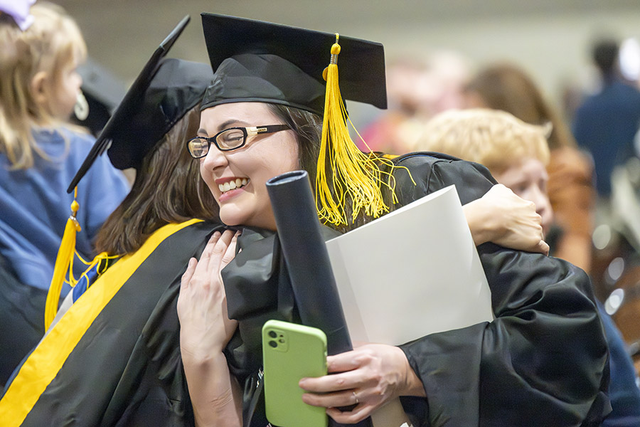 female with glasses wearing a graduation cap and gown gets a hug from a friend.