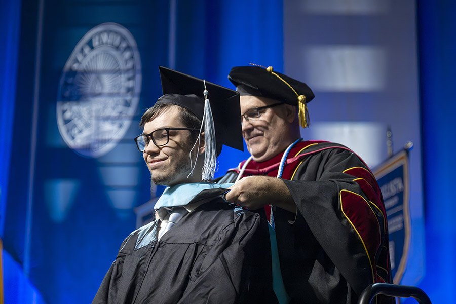 male student in a graduation cap and gown squatting to receive a hood