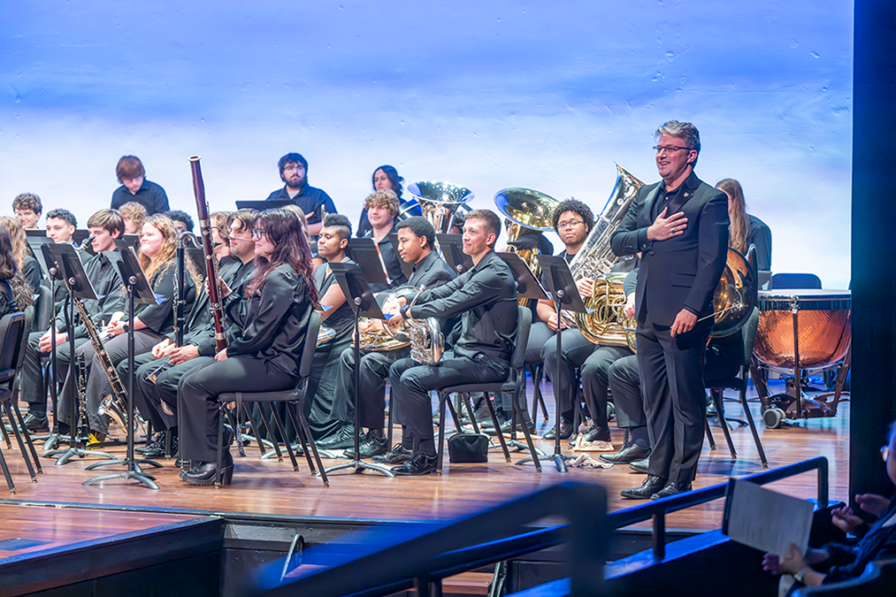 Man in black tie attire points stands with his hand over his heart in front of several rows of musicians holding various instruments.
