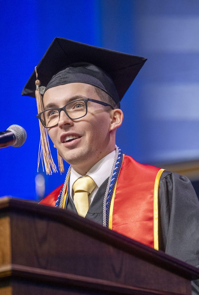 A male college graduate in full regalia stands at a podium and gives a speech to his fellow graduates.