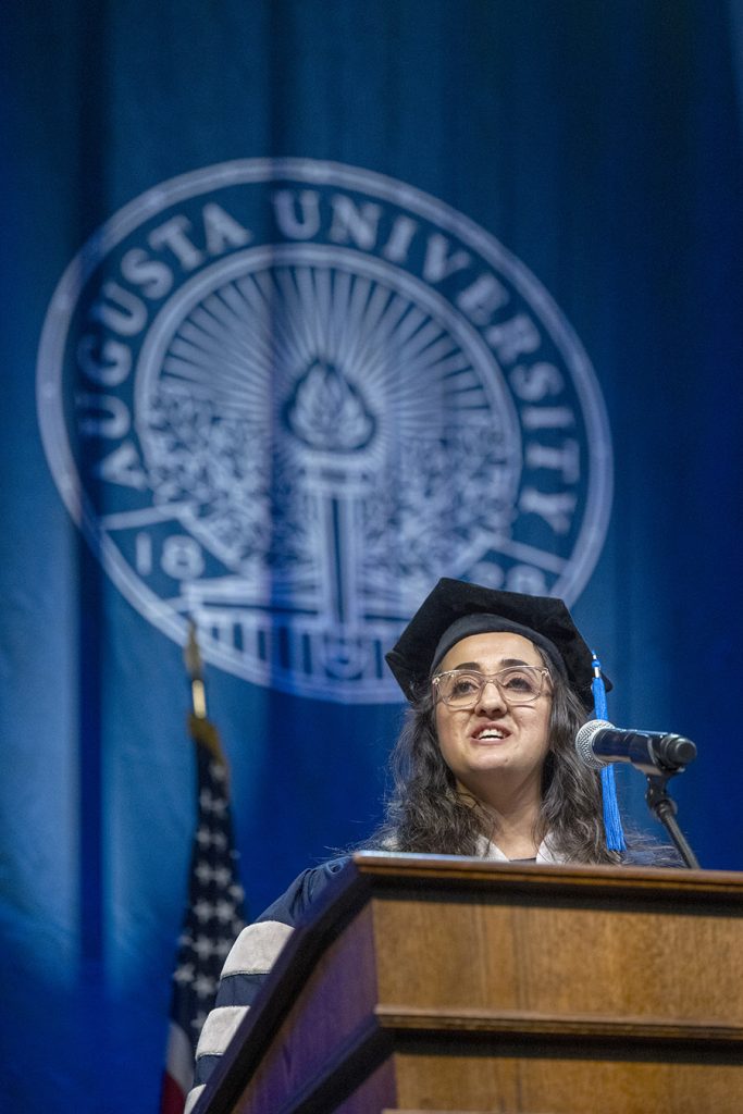 A female college graduate in full doctoral graduation regalia stands at a podium and speaks to her fellow college graduates.
