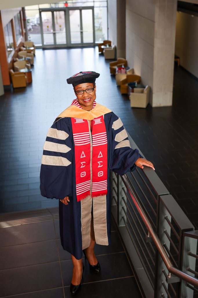 A woman wearing full graduation regalia for a Doctor of Nursing Practice candidate looks out of a window