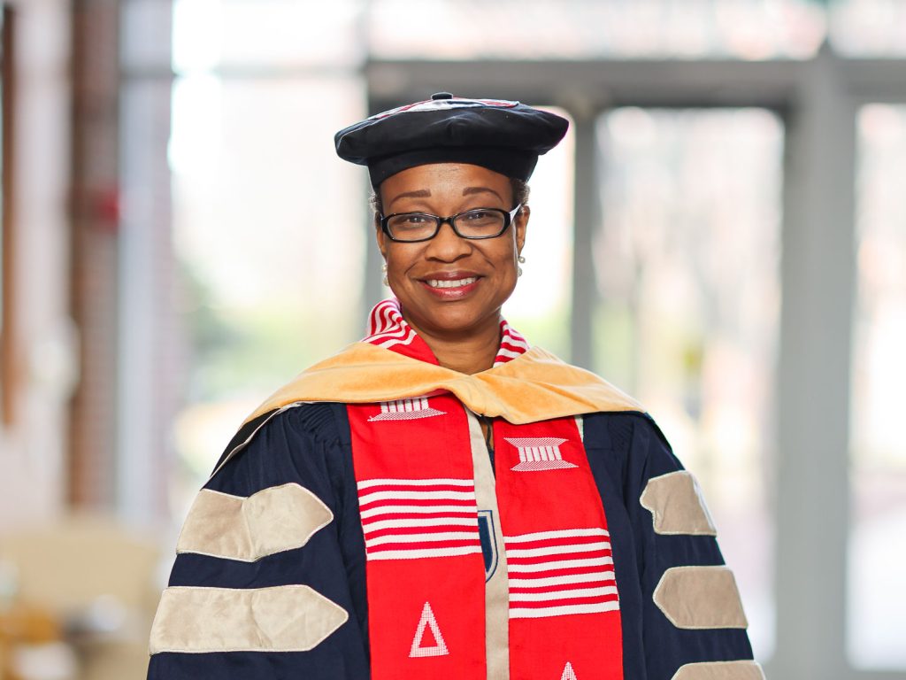 A woman wearing full graduation regalia for a DNP degree stands in front of window