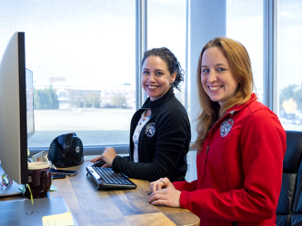 Two women smile, sitting in front of a window and computer monitor.