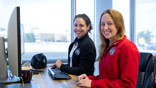 Two women smile, sitting in front of a window and computer monitor.