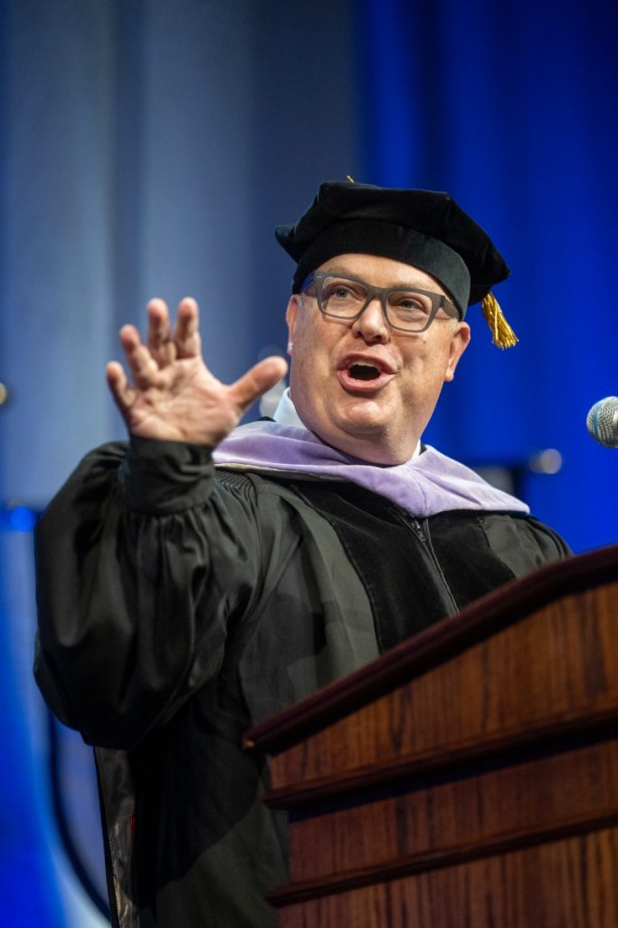 A man wearing full graduation regalia stands at a podium and speaks to a large group of college graduates and their families.