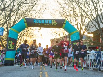 Runners begin a half marathon race at sunrise on the campus of a university. They are running under an inflatable arch with the logo for the Augusta Sports Council at the very top.