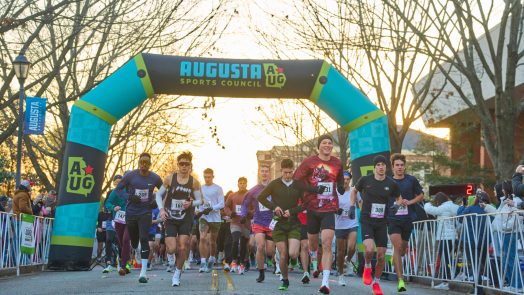 Runners begin a half marathon race at sunrise on the campus of a university. They are running under an inflatable arch with the logo for the Augusta Sports Council at the very top.
