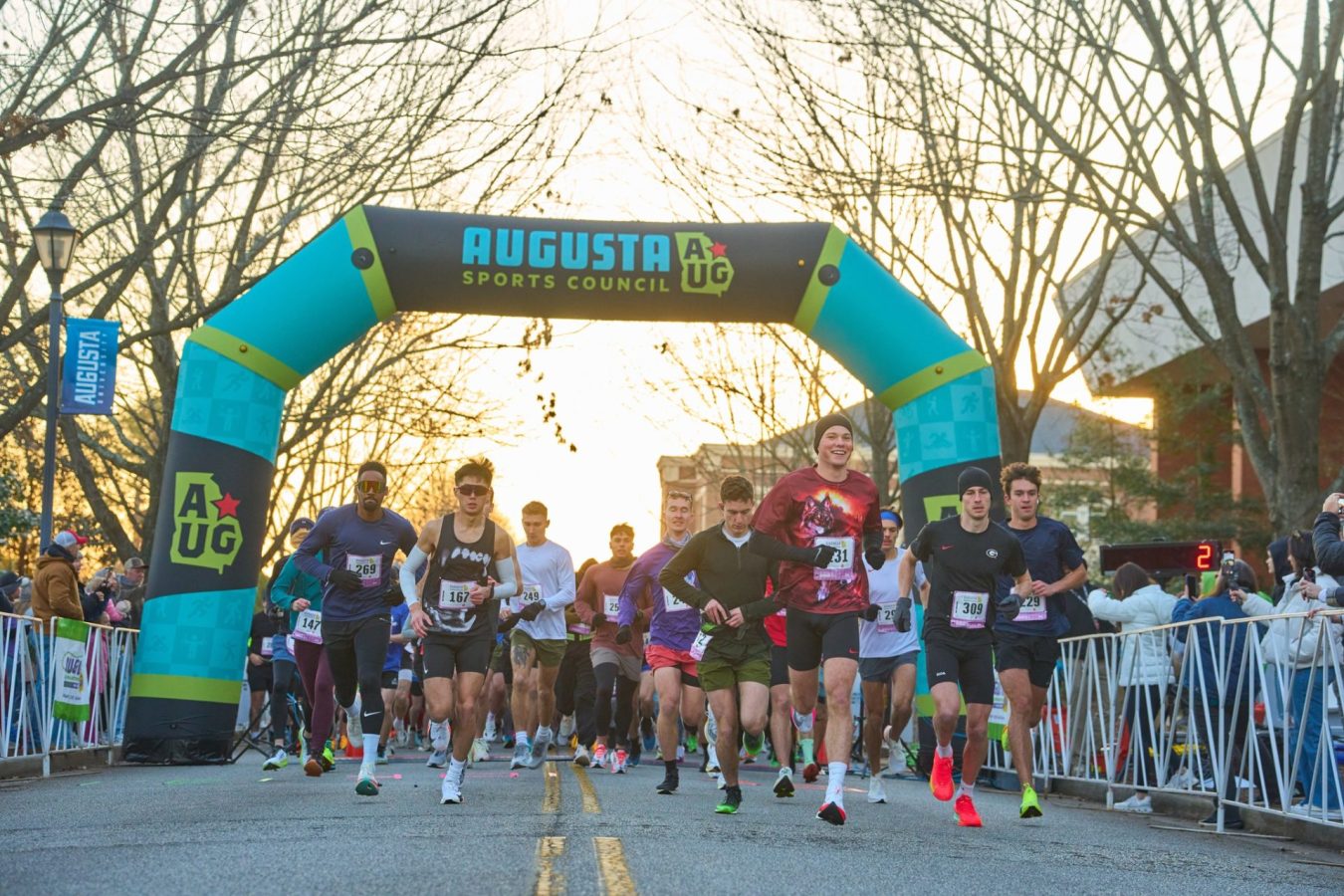 Runners begin a half marathon race at sunrise on the campus of a university. They are running under an inflatable arch with the logo for the Augusta Sports Council at the very top.