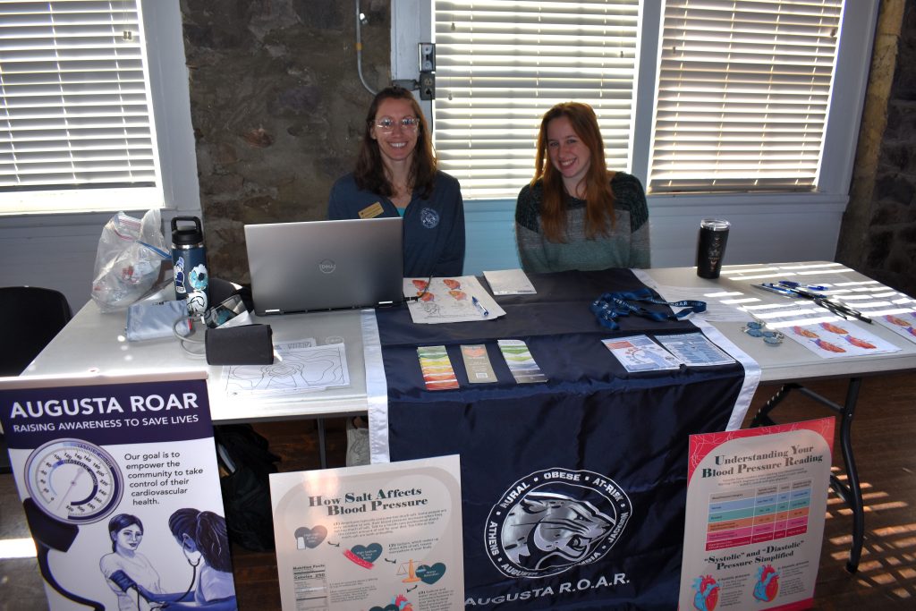 Two women sit behind a vendor table and smile at the camera