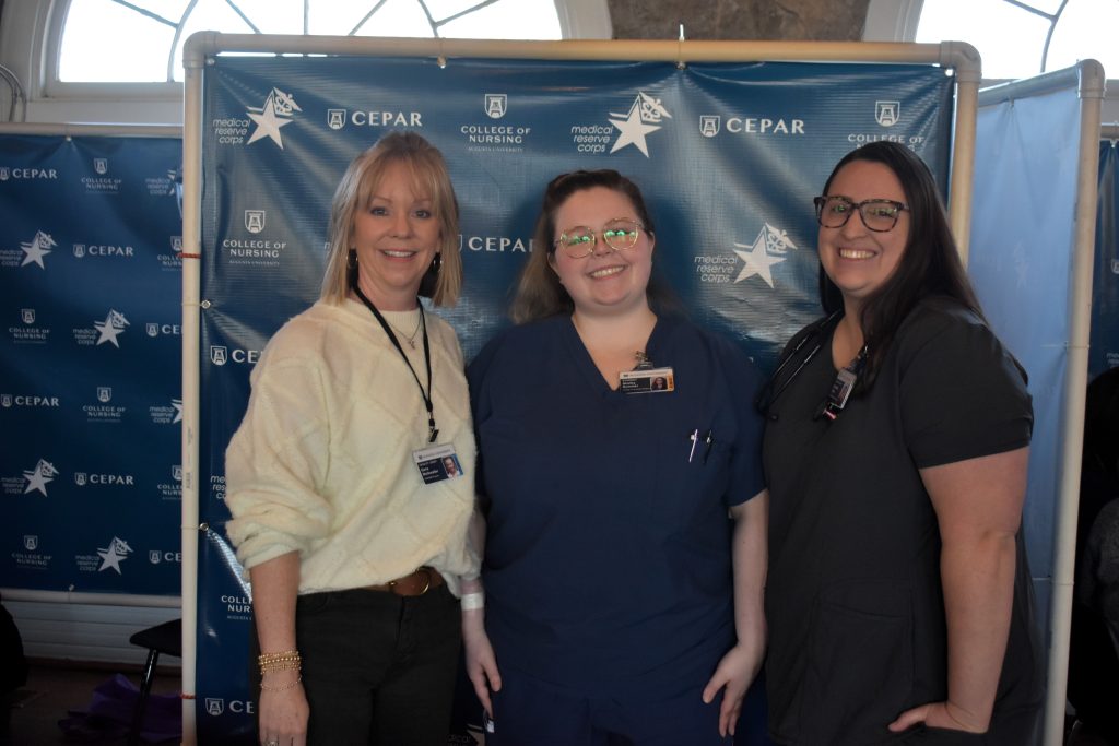 Three women, two wearing health care professional scrubs, stand inside a banquet room at a community center.