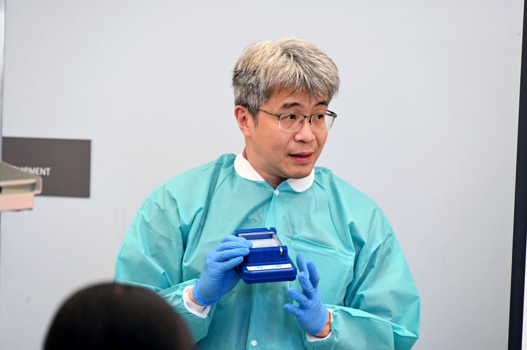 A male scientist in full lab gear speaks with a large group of high school students about the work that happens in his lab.