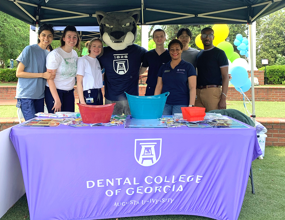 A group of adults stands under a tent in front of a table with someone wearing a large cat head for a costume.