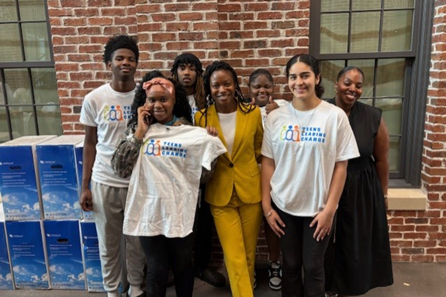 A group of five high school students and two women stand in a large atrium of a modern community building. They are helping hand out supplies to members of the community who are in need.