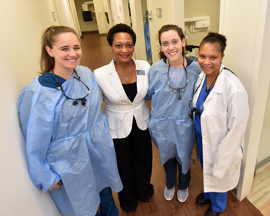 Four women stand in medical hallway, one in business dress and three in medical gear.