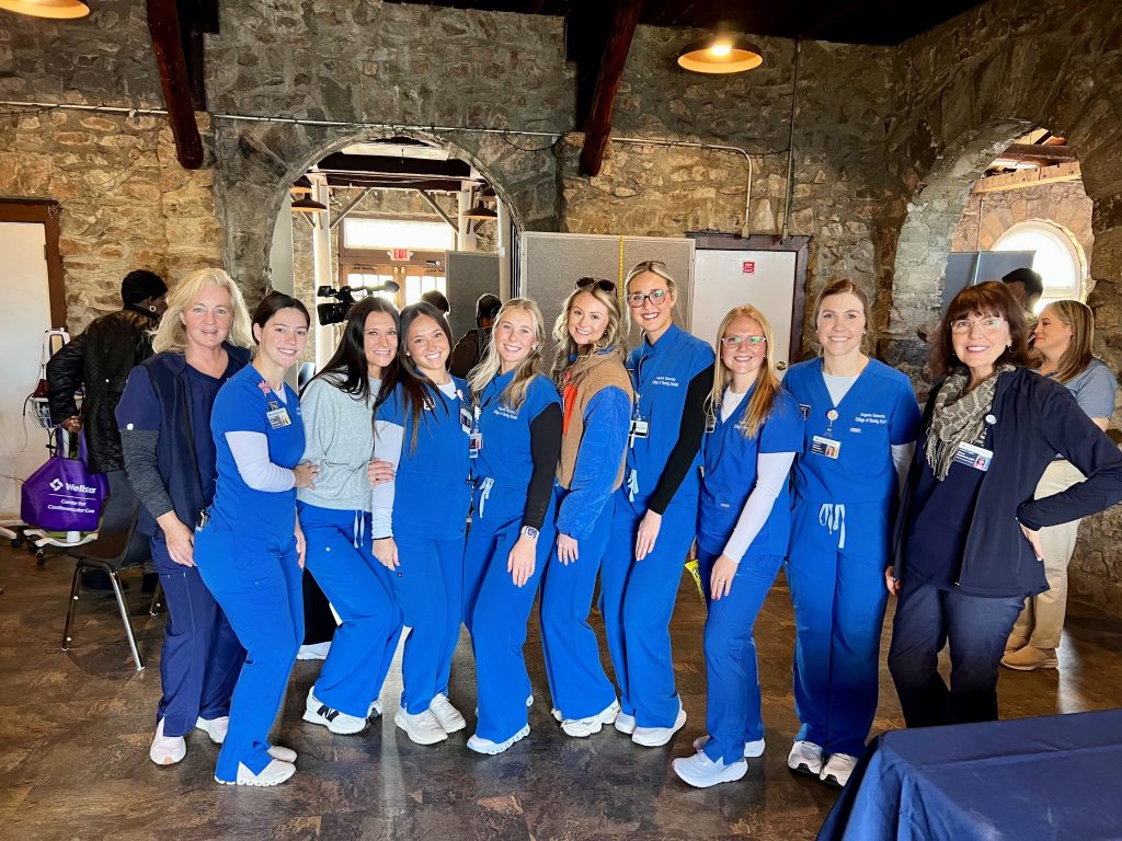 A group of 10 women, all wearing health care professional scrubs, stand inside a community center waiting to serve during a health clinic.