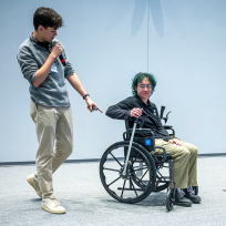 A female college student sits in a wheelchair with a special lever on each of the two bigger wheels. While she demonstrates how it works, a male college student uses a microphone to tell a large crowd how they came up with the idea to give people bound to a wheelchair more independence.