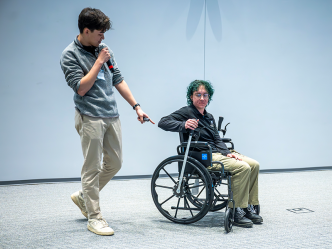 A female college student sits in a wheelchair with a special lever on each of the two bigger wheels. While she demonstrates how it works, a male college student uses a microphone to tell a large crowd how they came up with the idea to give people bound to a wheelchair more independence.