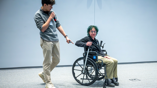 A female college student sits in a wheelchair with a special lever on each of the two bigger wheels. While she demonstrates how it works, a male college student uses a microphone to tell a large crowd how they came up with the idea to give people bound to a wheelchair more independence.