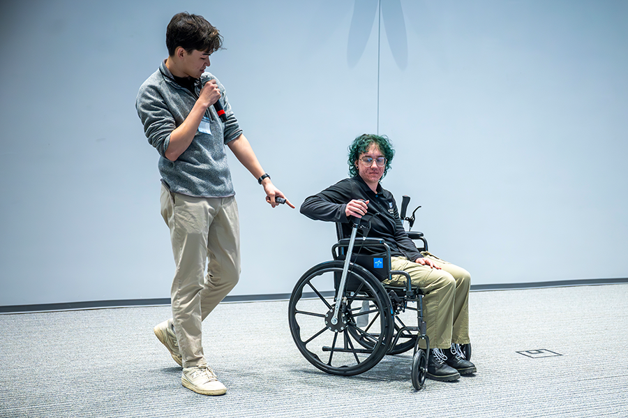 A female college student sits in a wheelchair with a special lever on each of the two bigger wheels. While she demonstrates how it works, a male college student uses a microphone to tell a large crowd how they came up with the idea to give people bound to a wheelchair more independence.