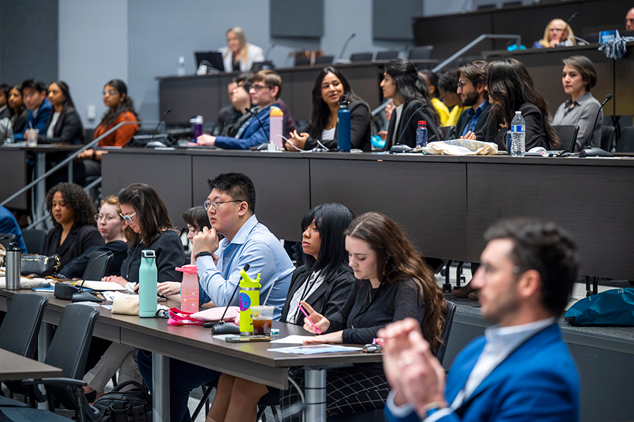 A group of college students sit in a large auditorium and listen to teams of students presenting ideas during a pitch competition.