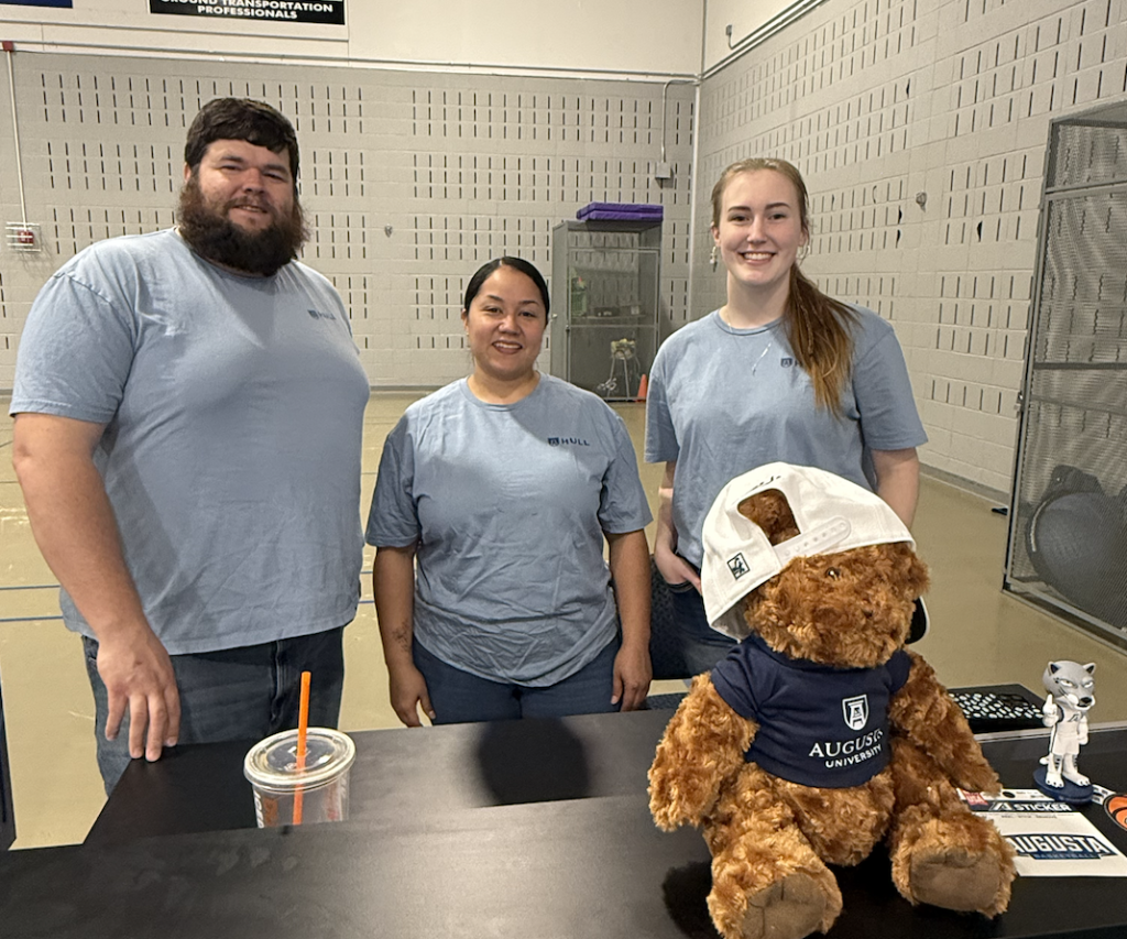 Three college students, two women and one man, stand behind a table and manage a merchandise booth during a sporting event.