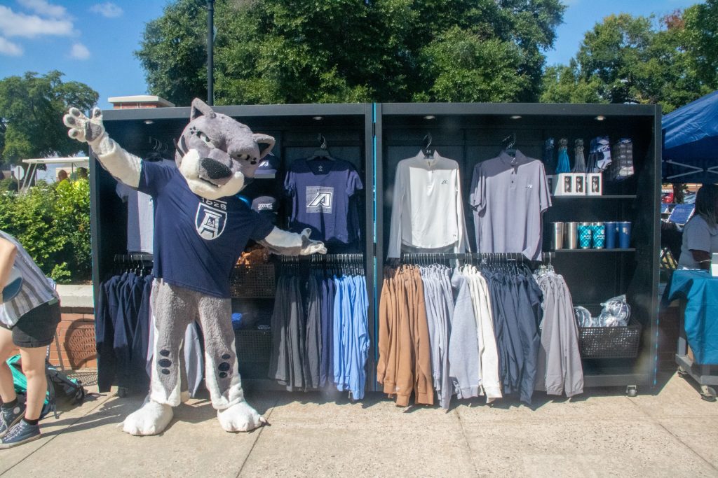A college mascot modeled after a large cat stands with a large cart of merchandise outside on a college campus.