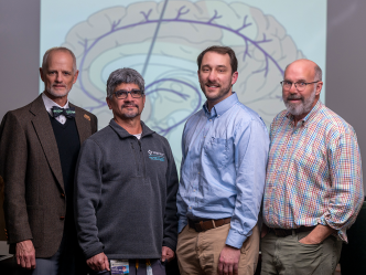 A group of four male neuroscientists stand in front of a backdrop with a picture of an animated brain on it and look at the camera.